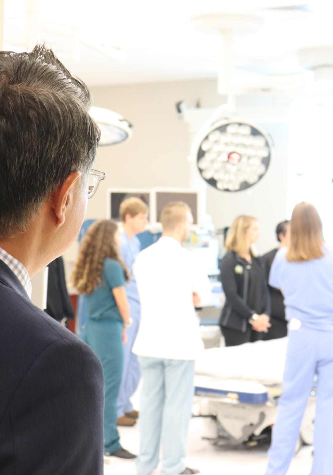 CAMLS A man in a suit is observing a group of medical professionals who are gathered around a patient bed in a hospital or clinical setting. The group includes individuals in scrubs and white coats, and there are medical lights and equipment in the background.