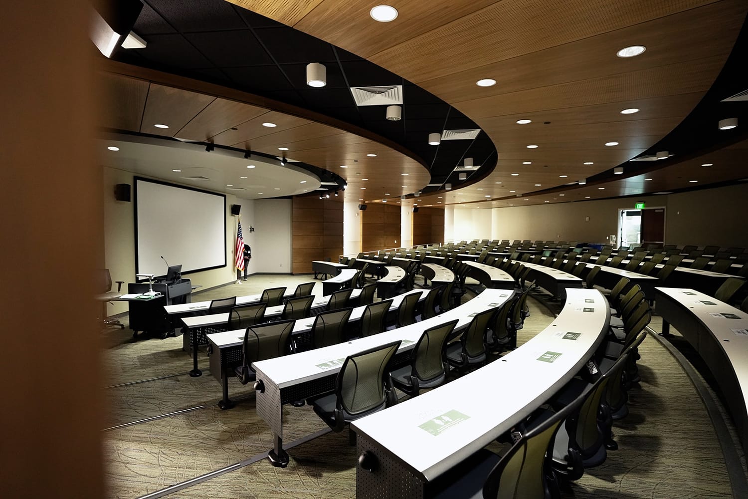 CAMLS A modern lecture hall with curved rows of desks and chairs, a large projection screen at the front, and an American flag in the corner. The room is well-lit with overhead lights and features wooden accents on the walls and ceiling. The desks have nameplates.