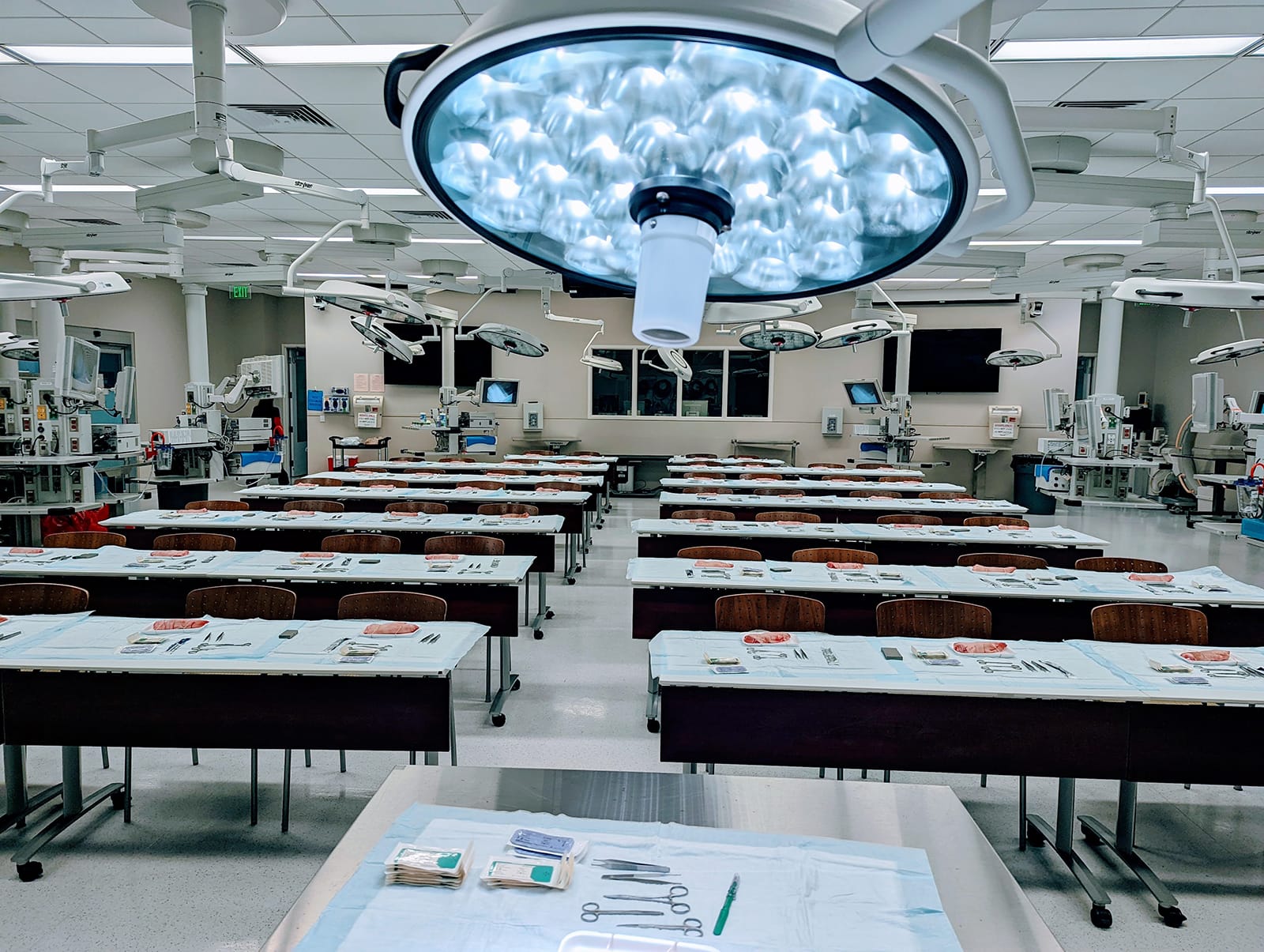 CAMLS A spacious, well-lit medical classroom with numerous tables set up for surgical training. Each table has a surgical drape and an array of medical instruments. Overhead surgical lights are positioned above each table, and advanced medical equipment is visible in the background.