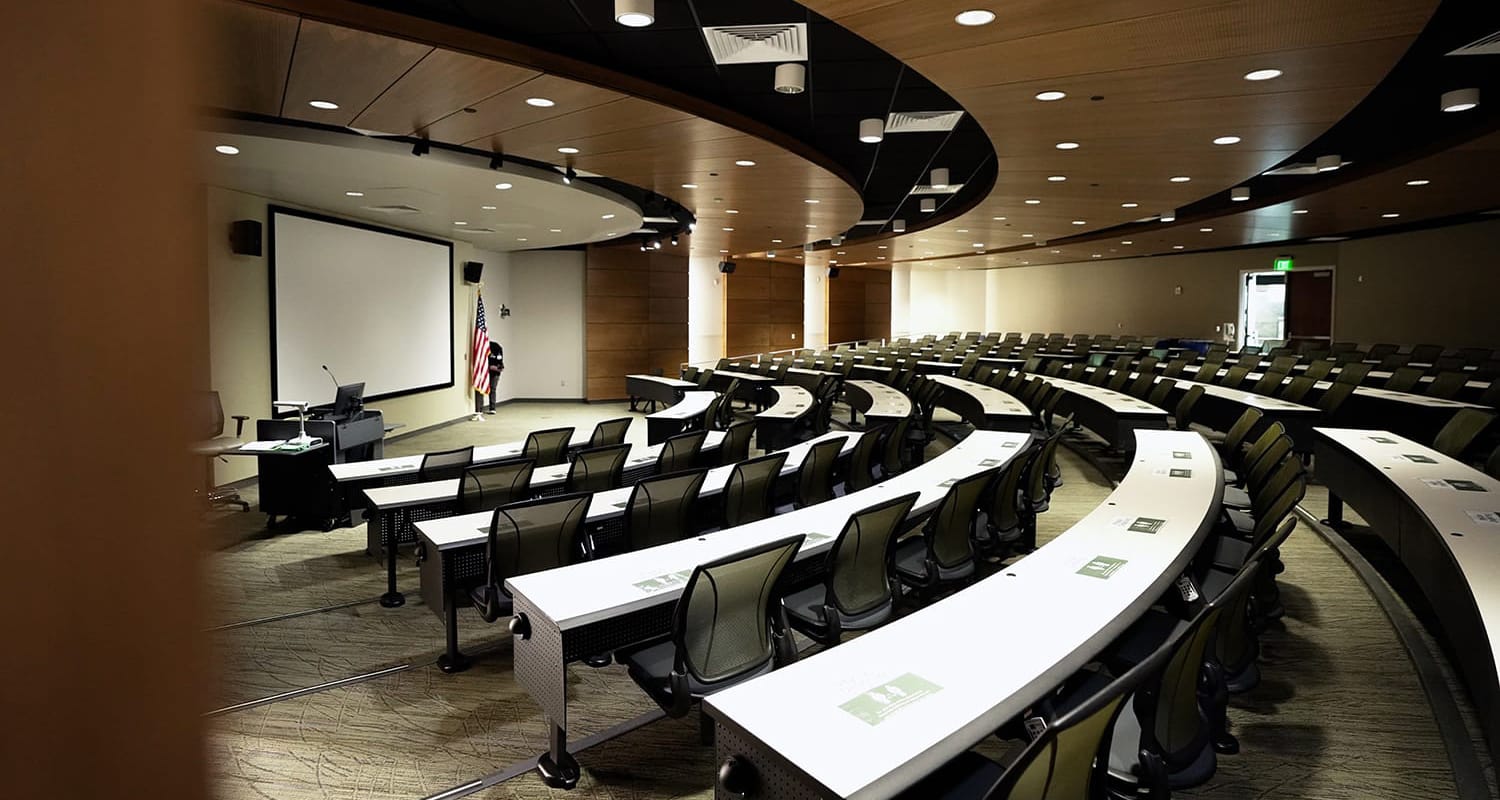 CAMLS A modern lecture hall with a curved, tiered seating arrangement facing a large projection screen at the front. Each desk has a chair and a book on it. The room features wooden walls and ceiling panels, with an American flag near the front podium.