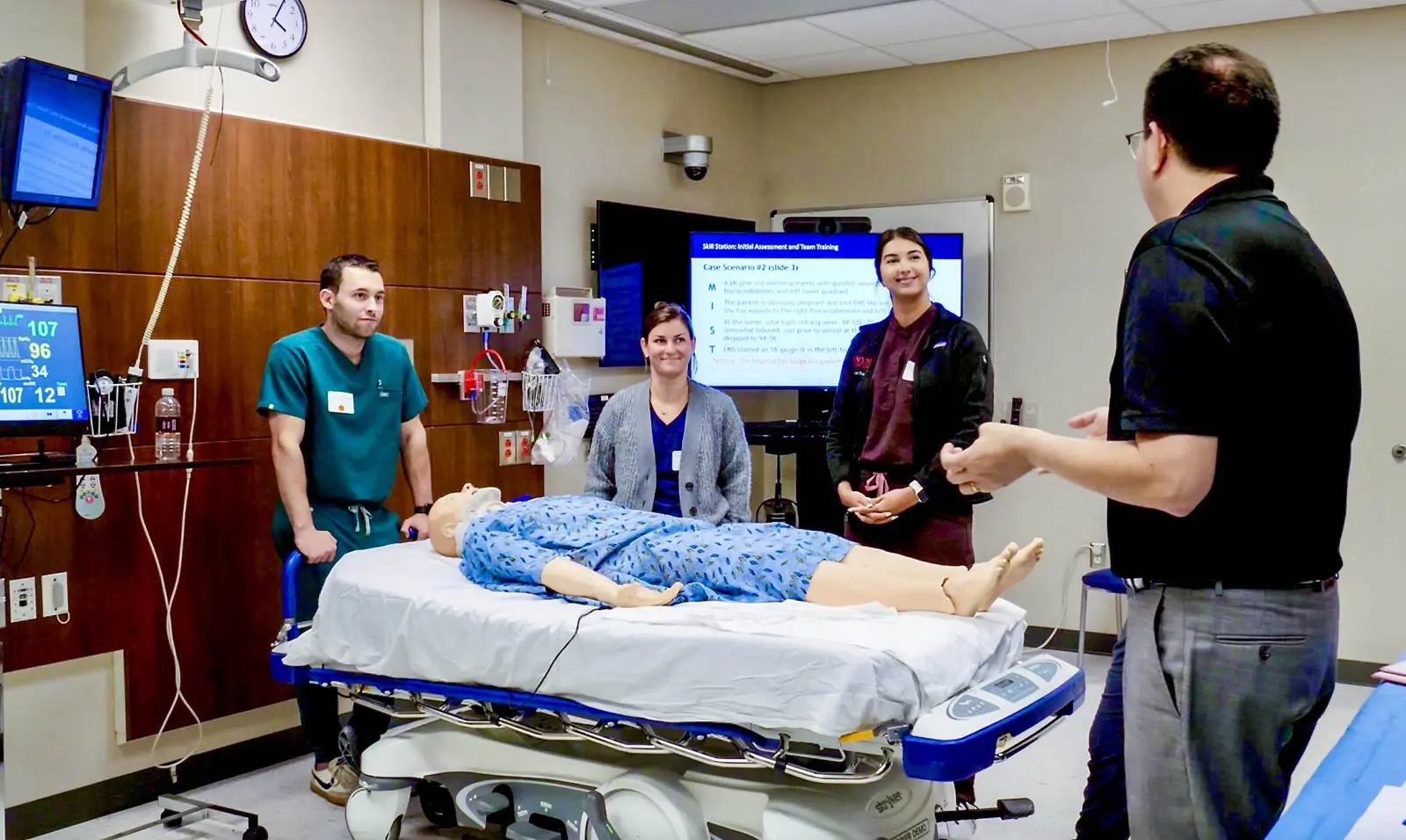 CAMLS A group of medical professionals stand around a hospital bed with a medical mannequin lying on it. A person is teaching from a screen displaying educational content. They are in a clinical setting, likely engaged in a training or simulation session.