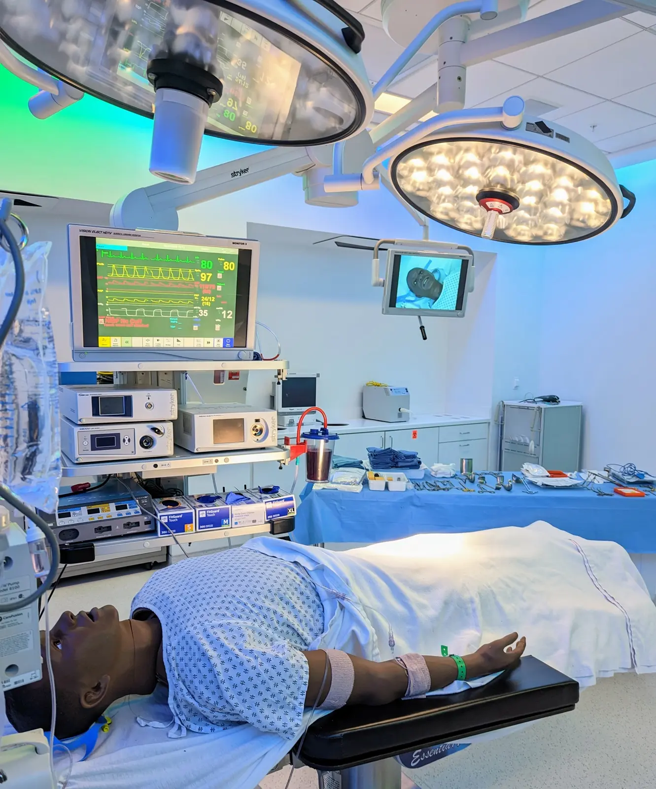 CAMLS A medical dummy lies on an operating table in a brightly lit operating room. Surrounding the dummy are various medical equipment and monitors displaying vital signs. Overhead surgical lights illuminate the scene, and a tray of surgical instruments is nearby.