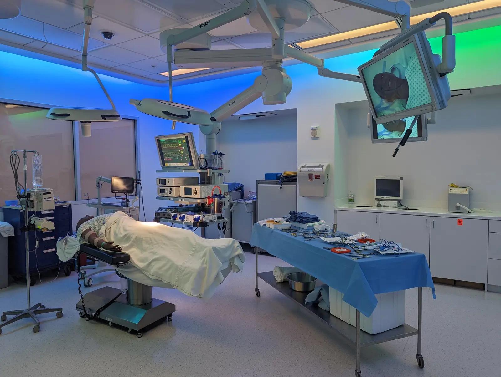 CAMLS A modern operating room with advanced medical equipment. An empty patient bed is covered with a white sheet. Monitors and surgical tools are arranged neatly. The lighting has a calming blue and green hue, and sterile draped tables are set up for a procedure.