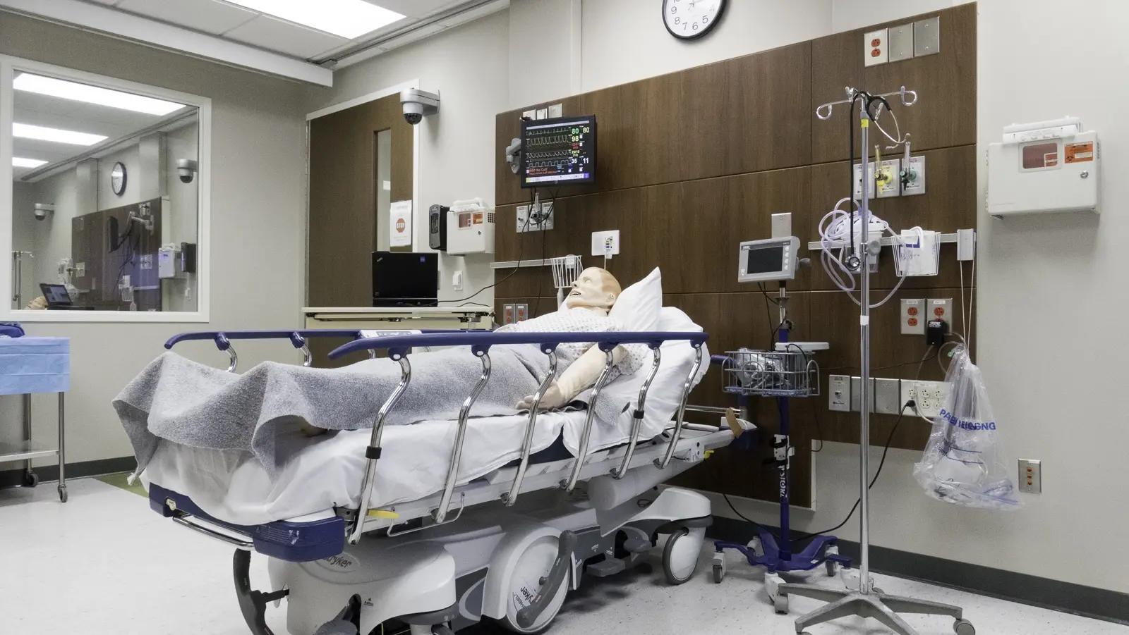 CAMLS A hospital room with a medical mannequin lying on a bed, covered with a gray blanket. The room features monitors, medical equipment, and an IV stand. The walls are equipped with various medical devices and a clock. The atmosphere is clinical and organized.