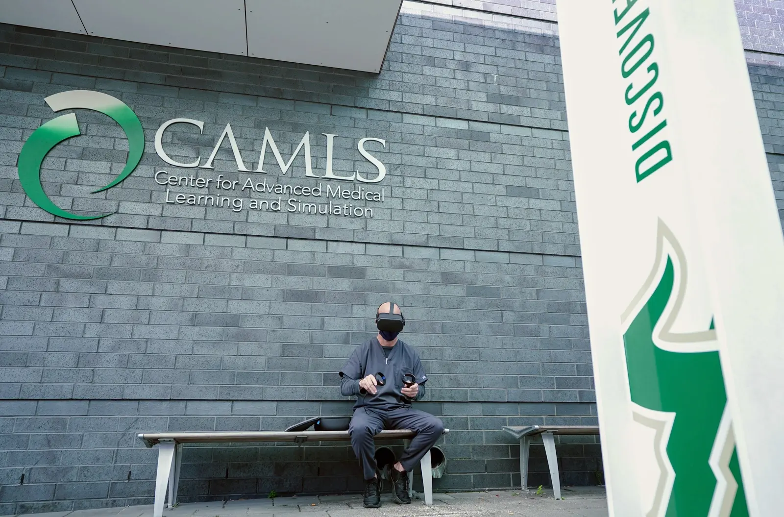 CAMLS A person wearing a virtual reality headset sits on a bench outside the CAMLS (Center for Advanced Medical Learning and Simulation) building. The individual holds a tablet, and a vertical banner with "DISCOVE" is partially visible on the right side of the image.