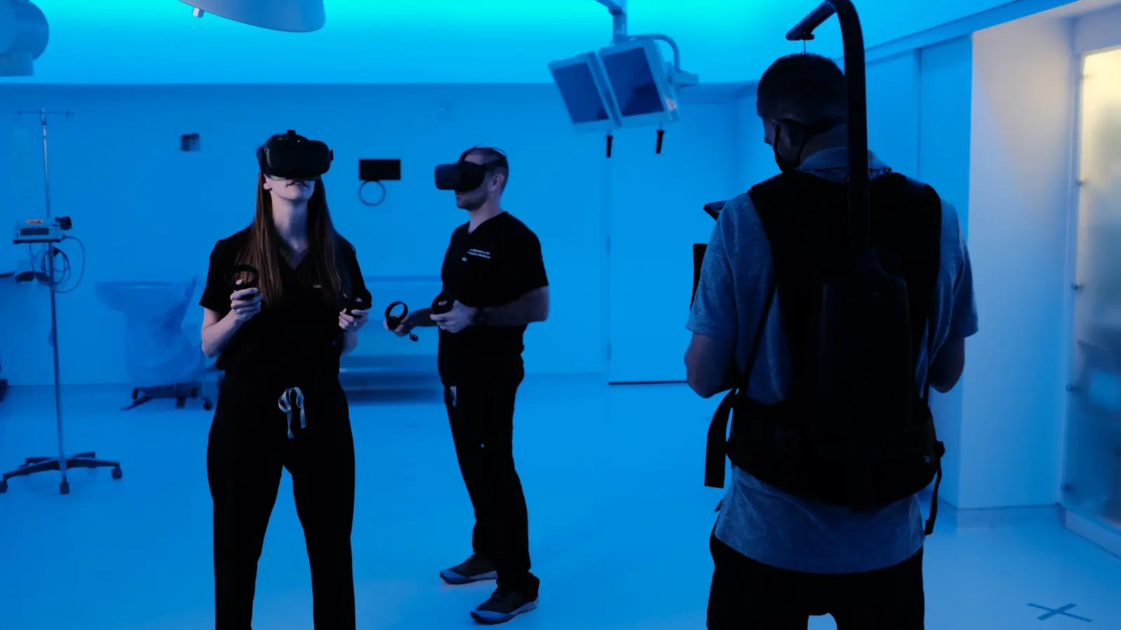 CAMLS Three people in a blue-lit room wear virtual reality headsets and hold controllers. They appear to be engaging with VR technology. One person has a strap across their back, possibly part of the VR equipment. The environment suggests a tech or training setup.