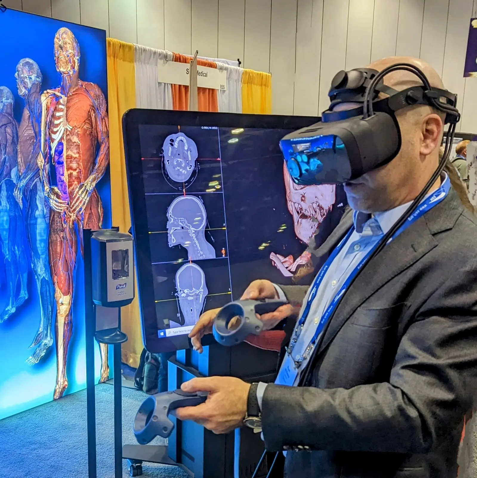 CAMLS A man wearing a suit and virtual reality headset uses VR controllers at a tech exhibition. Behind him, a large screen displays digital human anatomy images. An anatomical model with transparent layers is visible on a display board to the left.