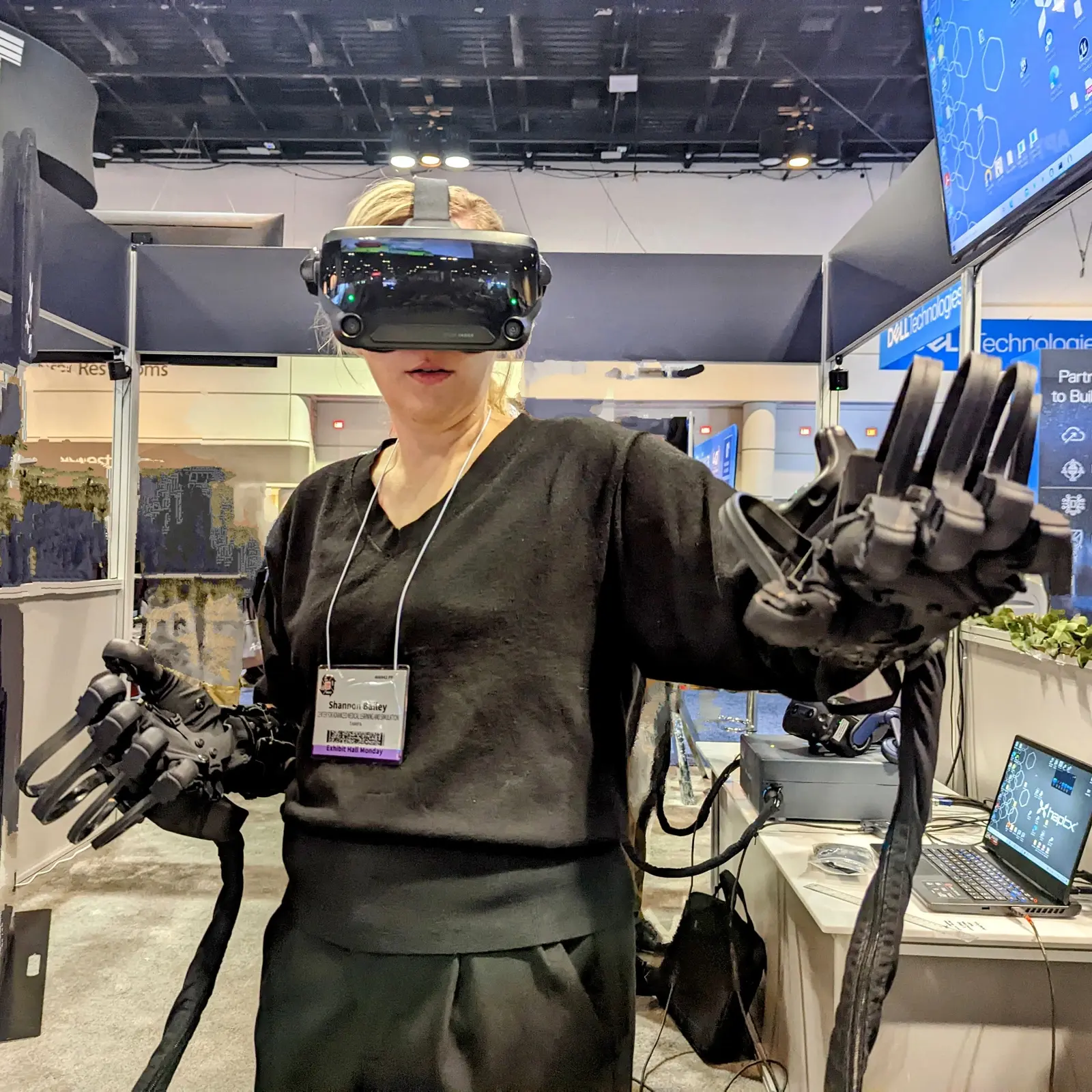 CAMLS A person is wearing a VR headset and haptic gloves, standing in a tech expo booth. They appear to be engaged in a virtual reality experience, with various electronic equipment and display monitors visible in the background.