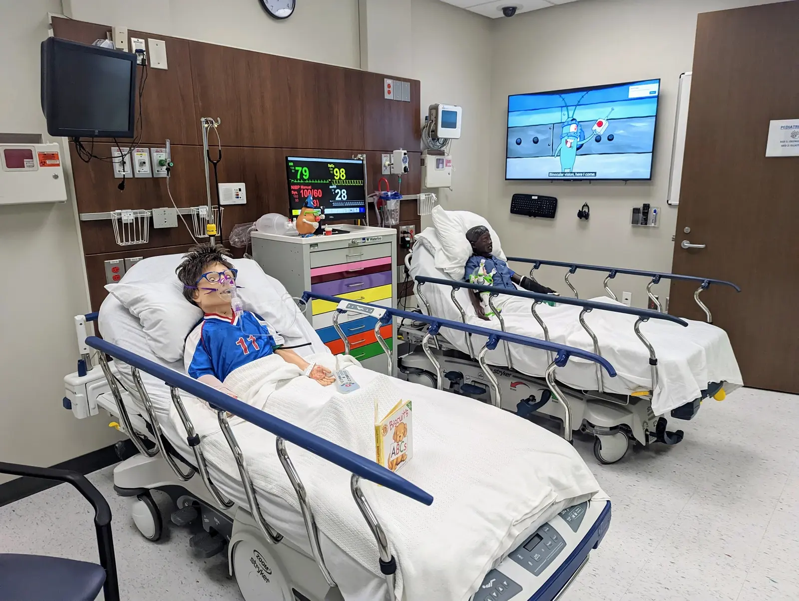 CAMLS Two children lie in hospital beds in a medical room, each connected to monitors displaying vital signs. Both children are watching a cartoon on a TV mounted on the wall. The room includes various medical equipment and storage cabinets.