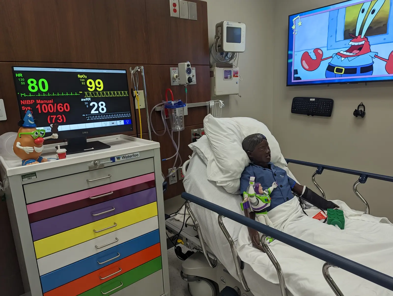 CAMLS A child is lying in a hospital bed, connected to medical monitors. A colorful set of drawers is beside the bed, and a monitor displays vital signs. The child is watching an animated cartoon on a wall-mounted TV. Medical equipment and cabinets are seen in the background.