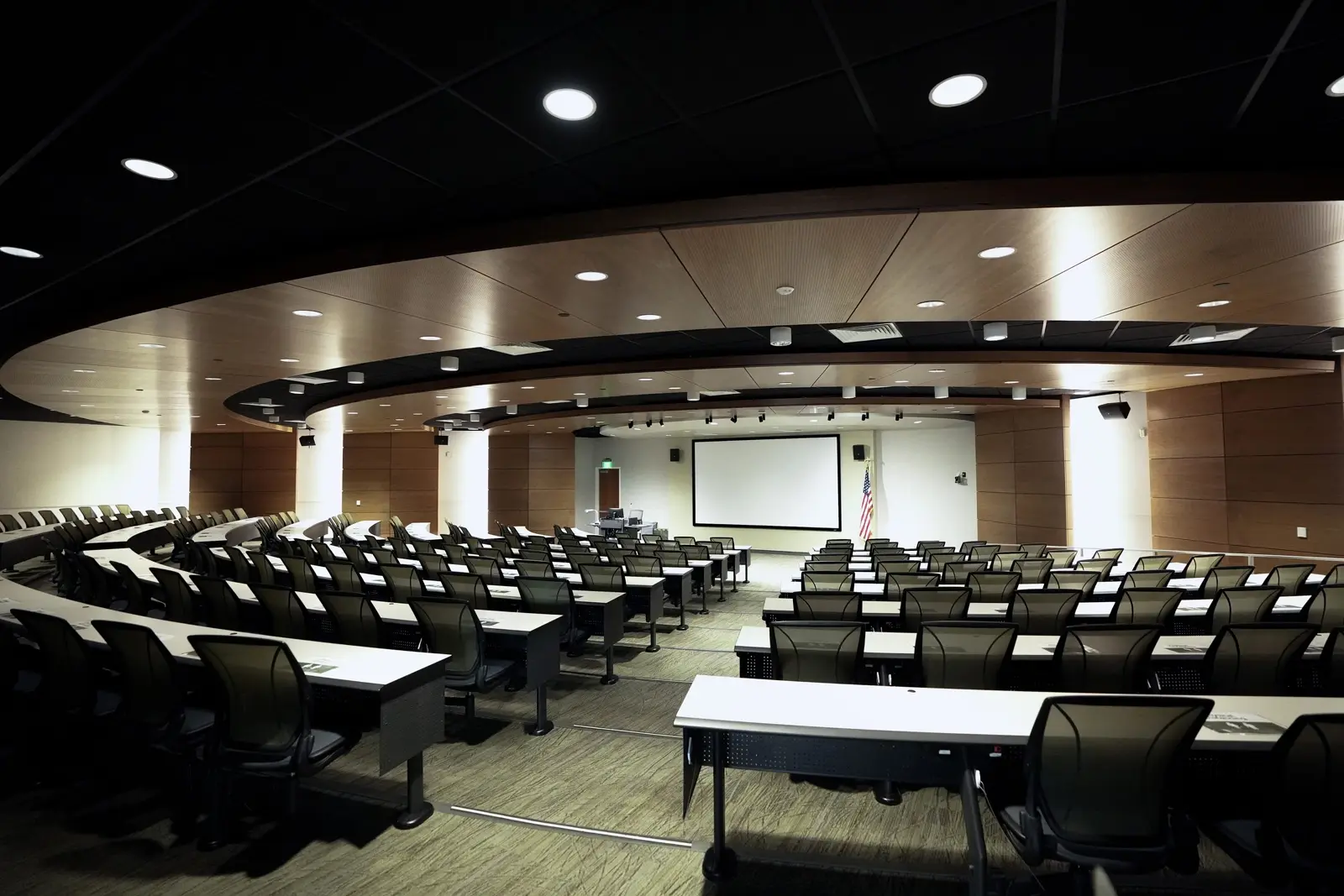 CAMLS A large, modern lecture hall with a curved layout, equipped with rows of desks and black mesh chairs. A projection screen, an American flag, and a podium are visible at the front. The ceiling features recessed lighting. The hall is empty.