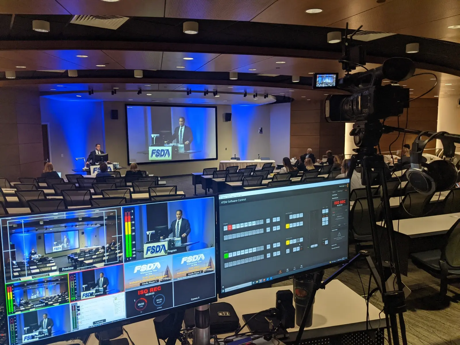 CAMLS Image of a conference room with a speaker presenting on stage to a seated audience. Multiple camera setups and a control station with monitors show the presentation being recorded or live-streamed. The speaker is displayed on a large screen behind them, surrounded by blue lights.