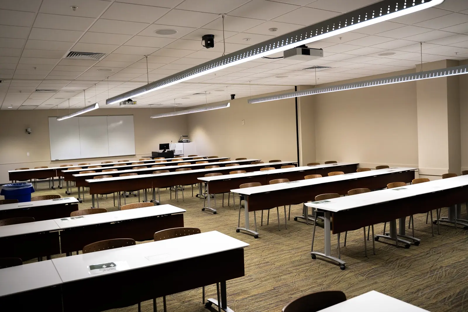CAMLS An empty classroom with rows of long desks and chairs facing a whiteboard and a podium. The ceiling has strip lighting, and a projector is mounted at the front. The walls and carpet are beige, creating a neutral tone throughout the space.