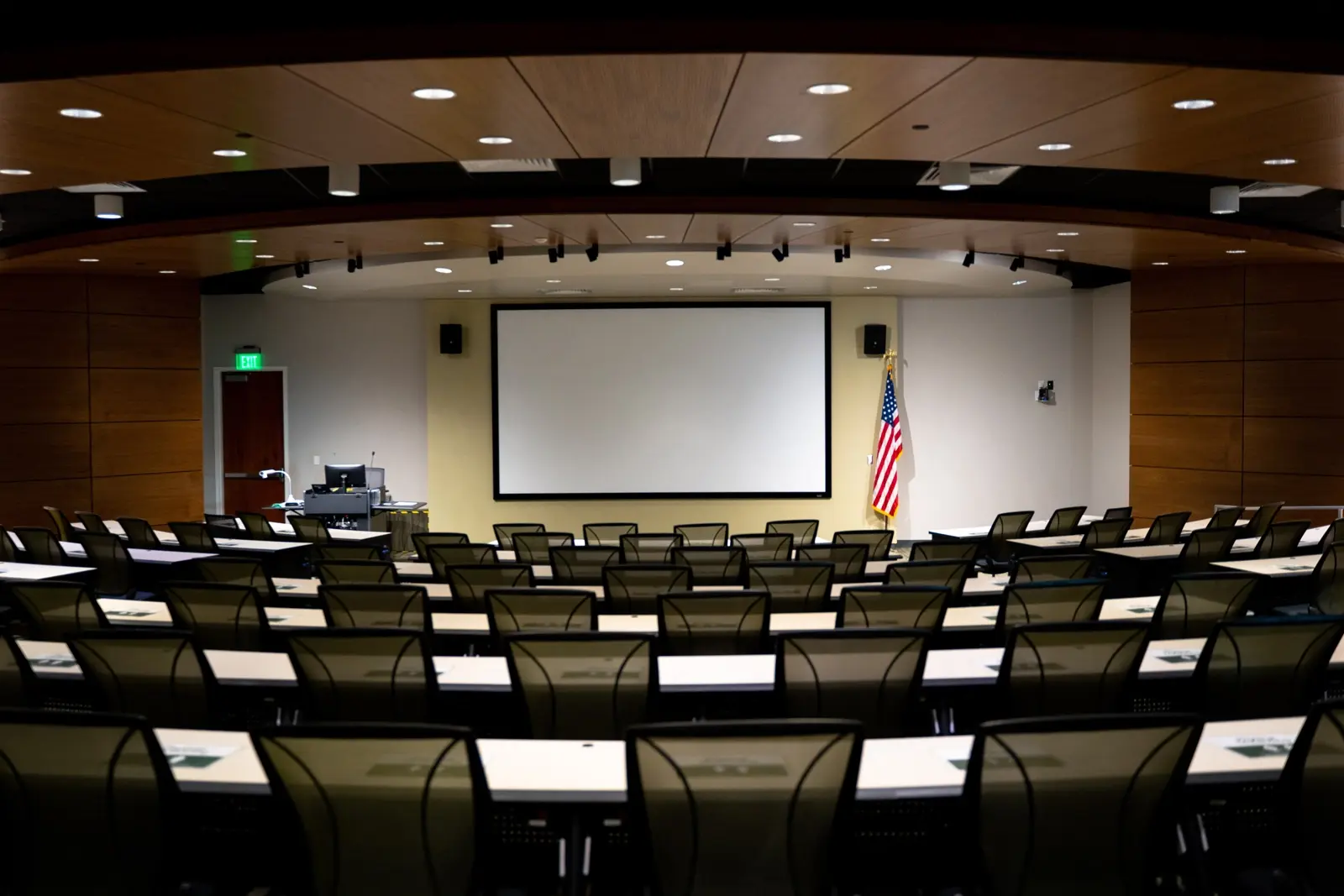 CAMLS A modern, empty lecture hall with rows of black chairs and white desks facing a large projection screen. The room features wooden paneling, ceiling lights, and an American flag standing to the side of the screen. A podium and equipment are also at the front.