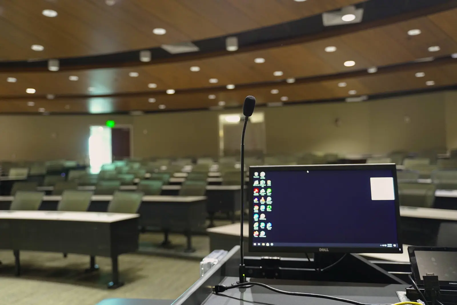CAMLS A lecture hall with rows of empty seats is pictured. In the foreground, there's a podium equipped with a microphone and a Dell monitor displaying a desktop with various icons. The hall's curved wooden ceiling and a partially visible green exit sign are also seen.