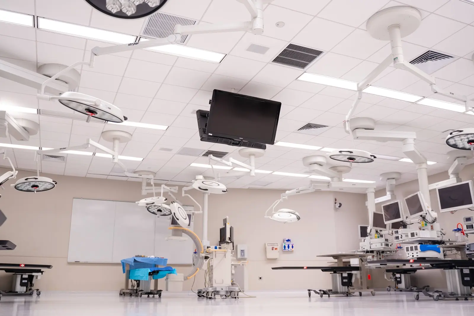 CAMLS A modern, clean, and brightly lit hospital operating room. The room features multiple surgical lights, flat-screen monitors, operating tables, and various advanced medical equipment. The walls are neutral in color, and there is a whiteboard in the background.