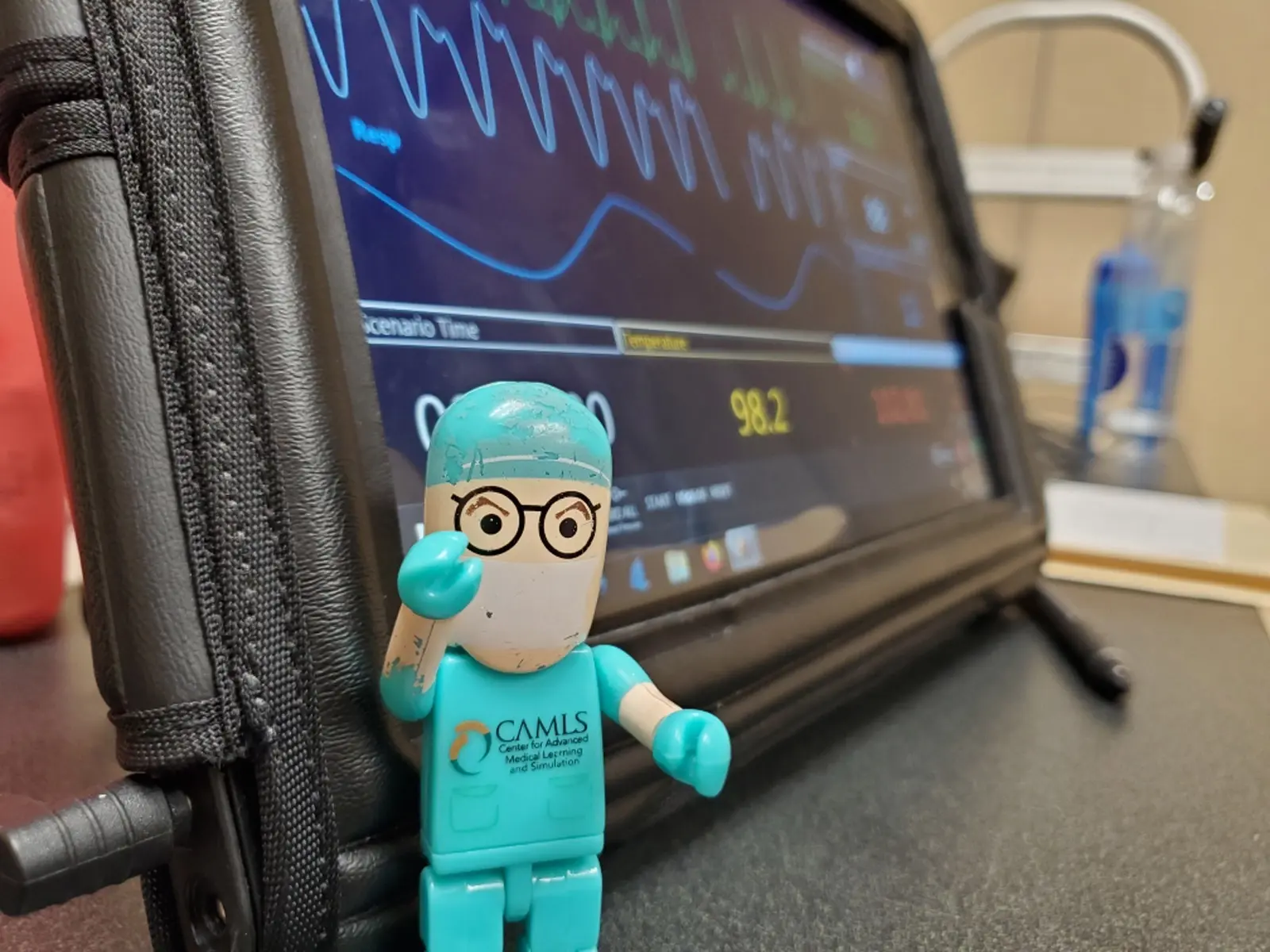 A small toy figurine wearing medical scrubs, glasses, and a surgical cap stands in front of a medical monitor displaying vital signs. The figurine has "CAMLS" printed on its shirt. The background shows a blurred medical setting.