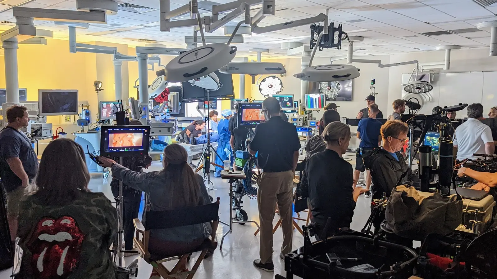 CAMLS A busy film crew is shooting a scene in a brightly-lit room filled with medical equipment, with multiple cameras and lighting rigs in use. Crew members are seen operating equipment, monitoring screens, and discussing the shot.