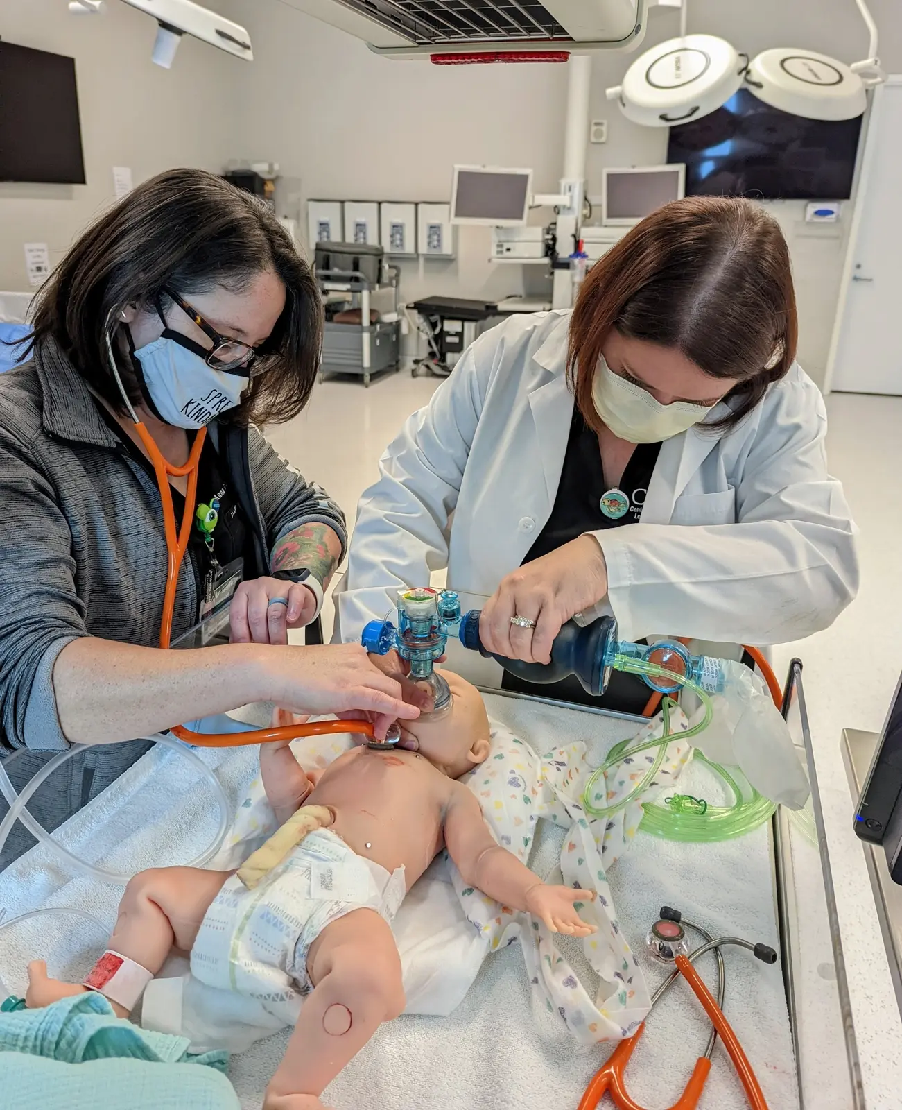 CAMLS Two healthcare professionals, wearing masks and gloves, are attending to a medical training mannequin of a baby. One is performing ventilation with a bag valve mask while the other assists, in a clinical setting with various medical equipment in the background.