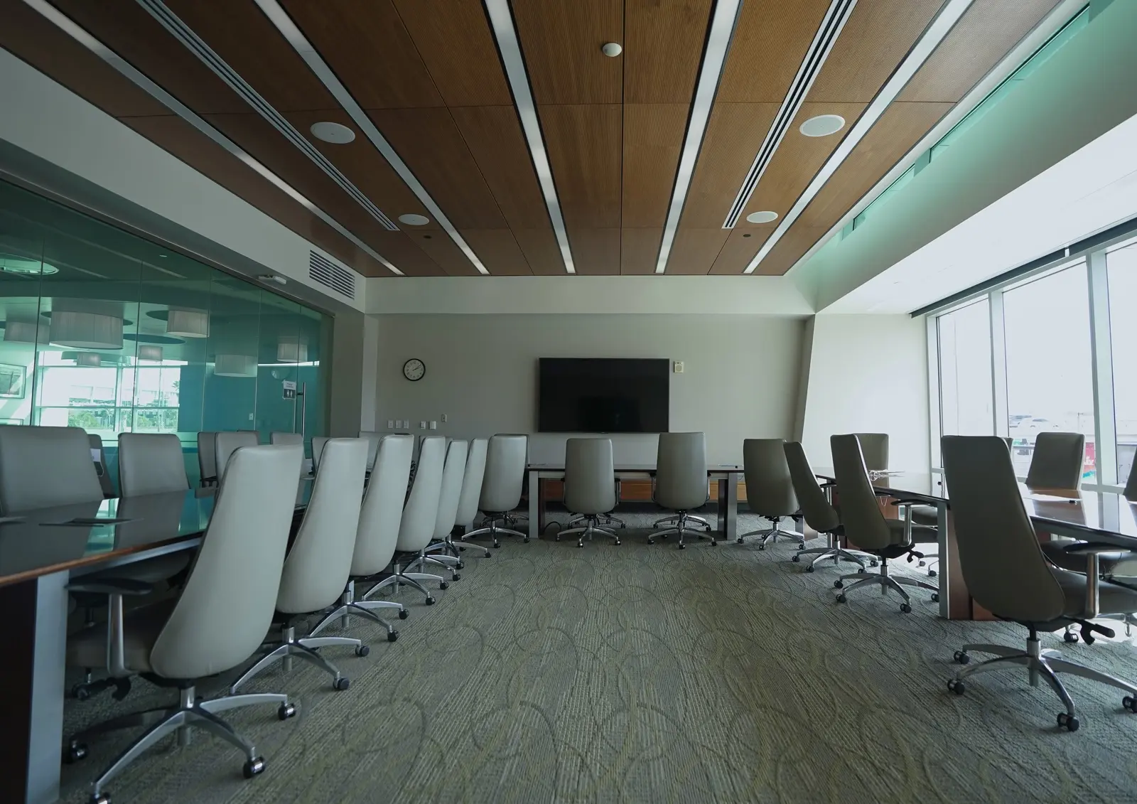 CAMLS A modern conference room with a large table surrounded by several high-backed office chairs. The room has a wooden ceiling with recessed lighting, a flat-screen TV on the wall, glass walls on one side, and large windows allowing natural light to fill the space.