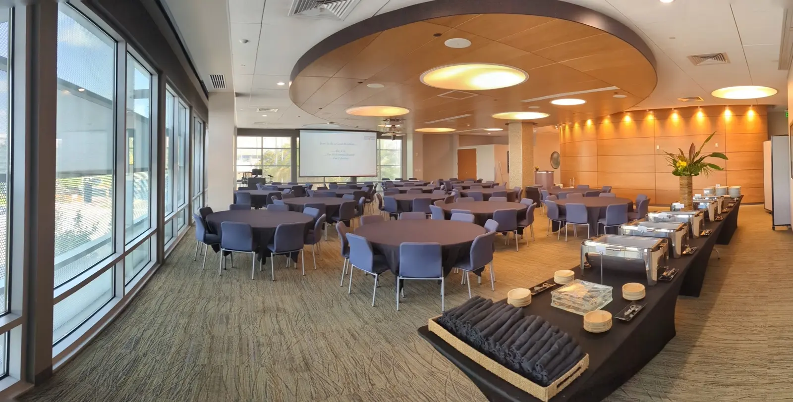CAMLS A modern conference room with large windows, circular chandeliers, round tables with blue chairs, and a buffet table to the right. A projector screen is visible at the front of the room. The setup is ready for an event or meeting.