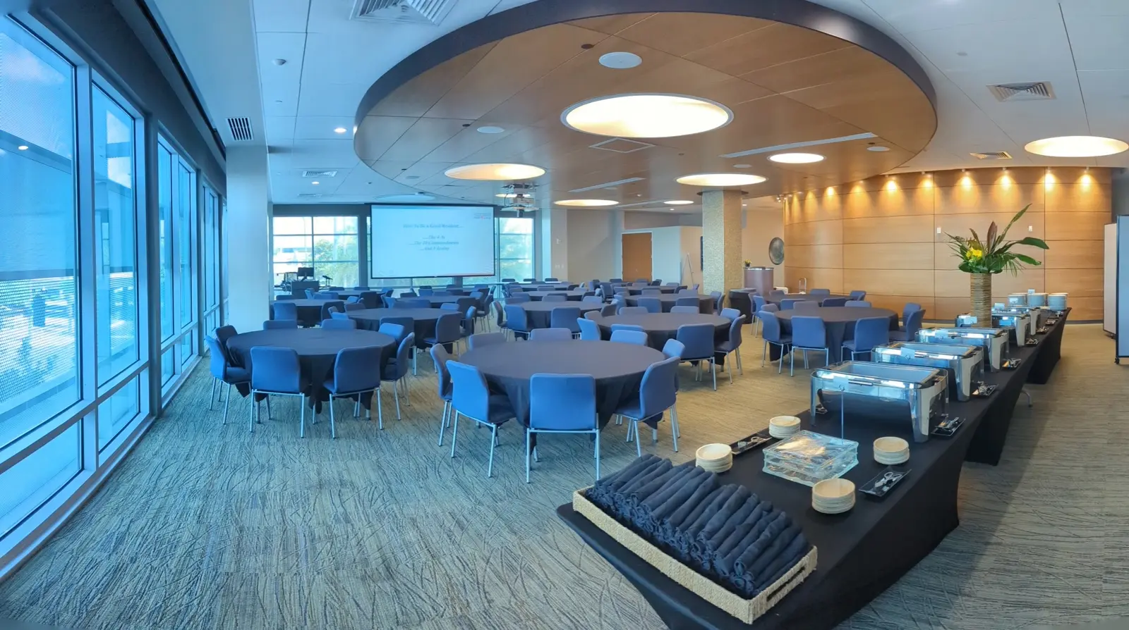 CAMLS A modern conference room with round tables covered in blue tablecloths and matching blue chairs. A buffet table with chafing dishes and neatly arranged utensils is set up on the right. Large windows let in natural light, and a projector screen is at the far end.