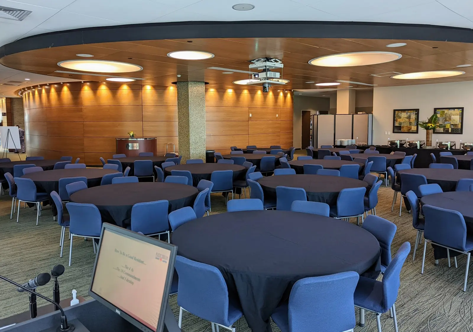 CAMLS A modern conference room with round tables covered in black tablecloths surrounded by blue chairs. In the background, there is a wooden wall with indirect lighting and a small reception desk. To the right, a buffet table is set up with refreshments.