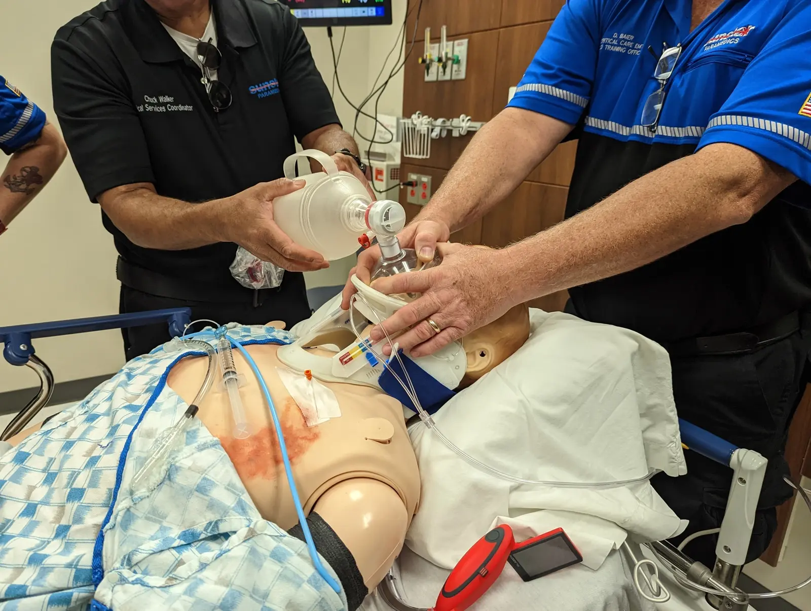 CAMLS Medical professionals demonstrate CPR techniques on a simulation mannequin. They use a bag valve mask for ventilation while monitoring hooked-up equipment. The scenario takes place in a clinical setting, with medical supplies and monitoring screens visible.