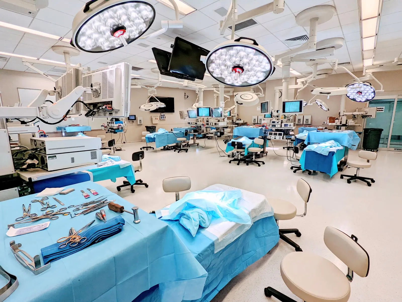 CAMLS A brightly lit surgical room equipped with multiple operating tables covered with blue drapes. Overhead surgical lights, monitors, and various medical instruments are neatly arranged. The room is clean and well-organized, ready for medical procedures.