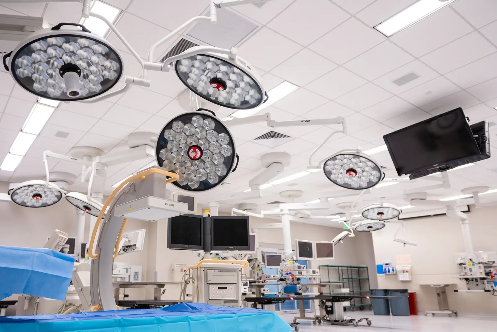 CAMLS A modern operating room equipped with several large round surgical lights on the ceiling, multiple medical monitors, and various surgical equipment. The room is well-lit with clean, sterile surfaces, and a blue draped surgical area is visible in the foreground.