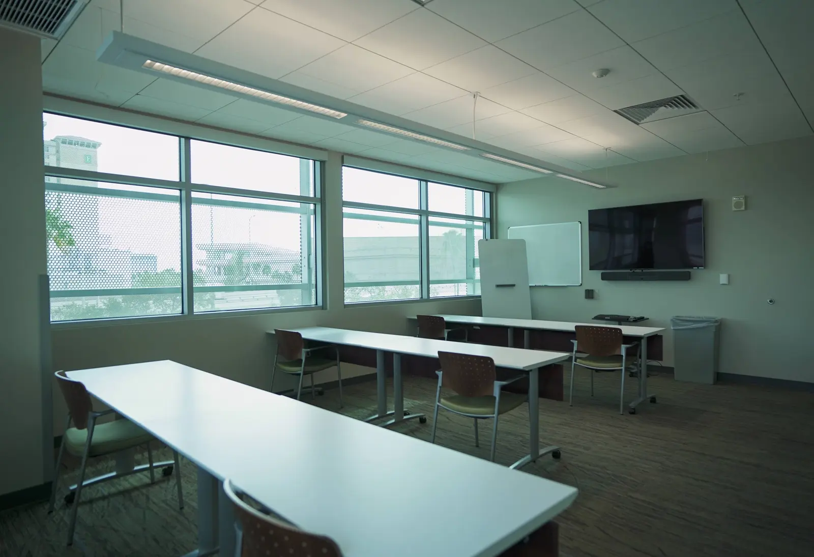CAMLS A brightly lit classroom with large windows, three white tables, and several chairs. The room has a wall-mounted flat-screen TV, a whiteboard, and light green walls. The ceiling lights are on, and the outside view includes buildings and a cloudy sky.