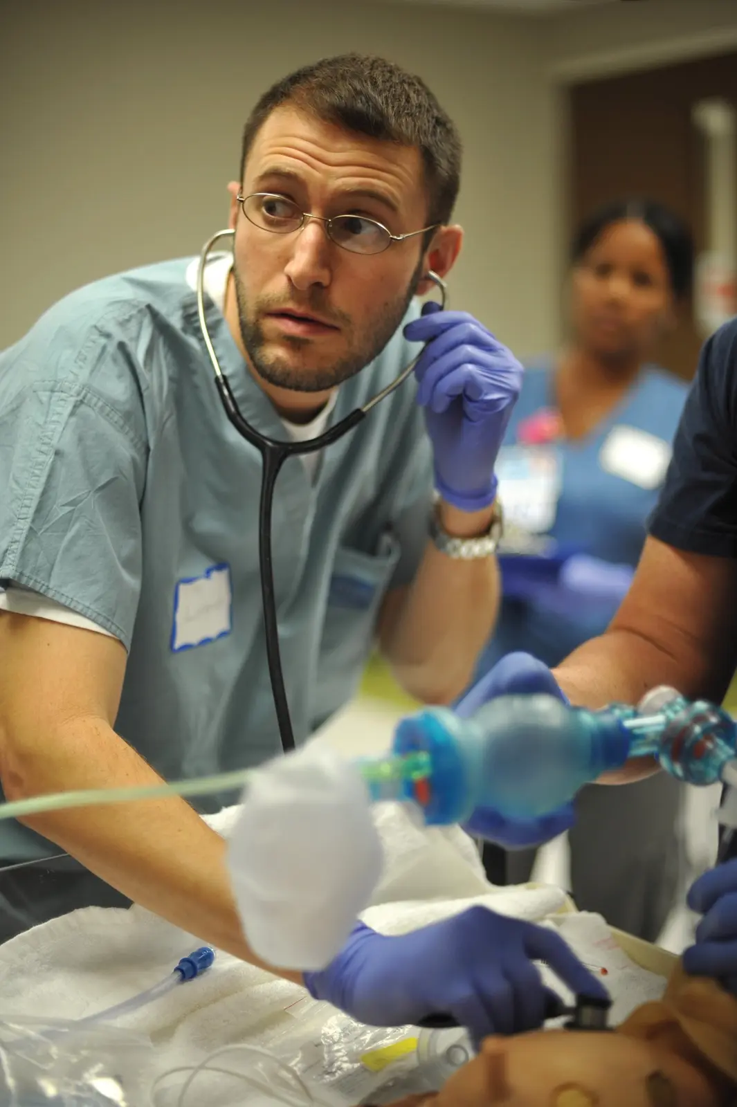 CAMLS A medical professional wearing scrubs, gloves, and a stethoscope listens attentively with the stethoscope to a simulated patient during a medical training session. In the background, other medical staff are visible, blurred, focusing on the training activity.