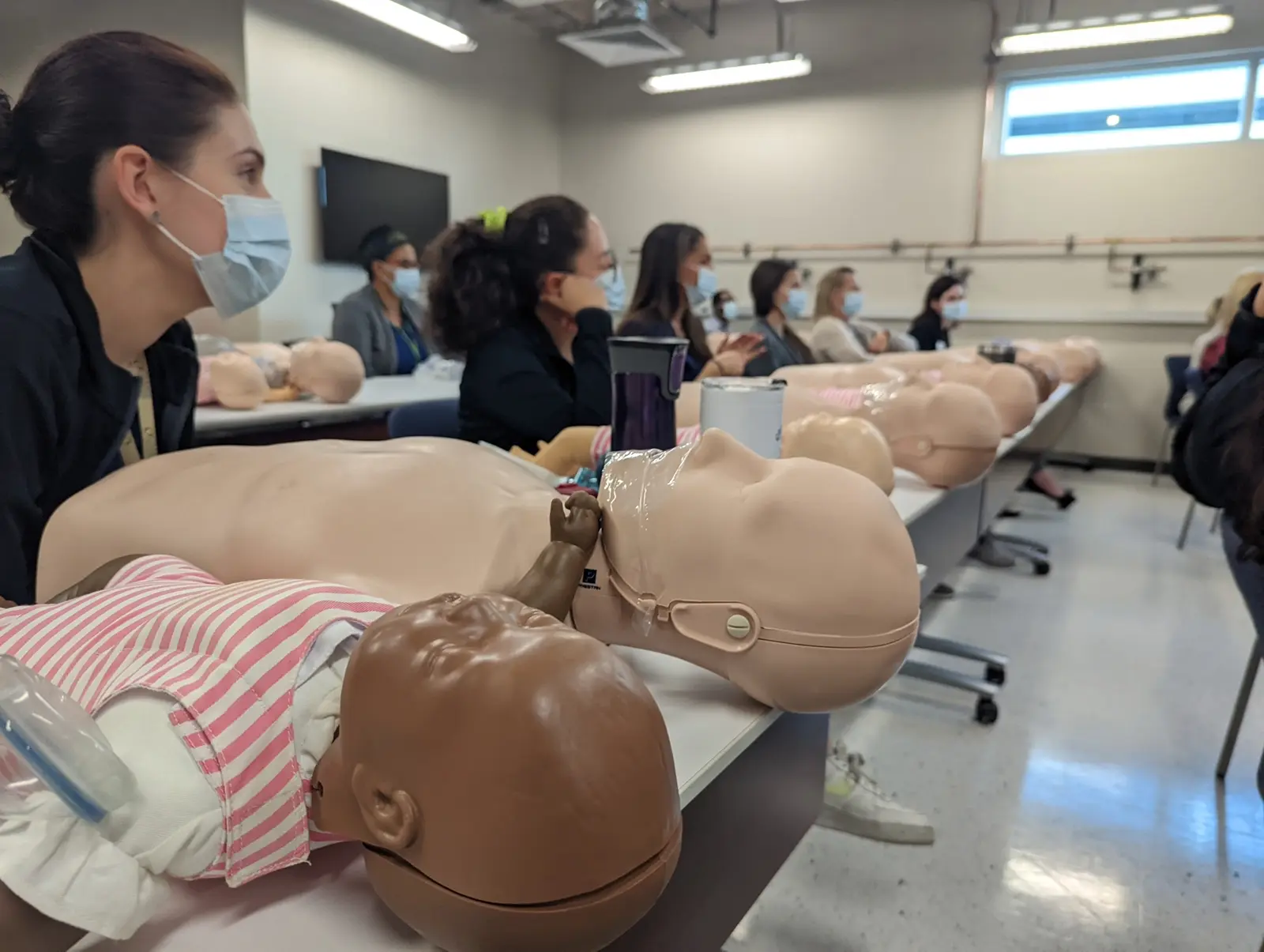 CAMLS A group of people wearing masks are seated at tables, attentively observing a training session. Mannequins of infants and adults are placed on the tables in front of them, suggesting that the session is related to CPR or medical training.