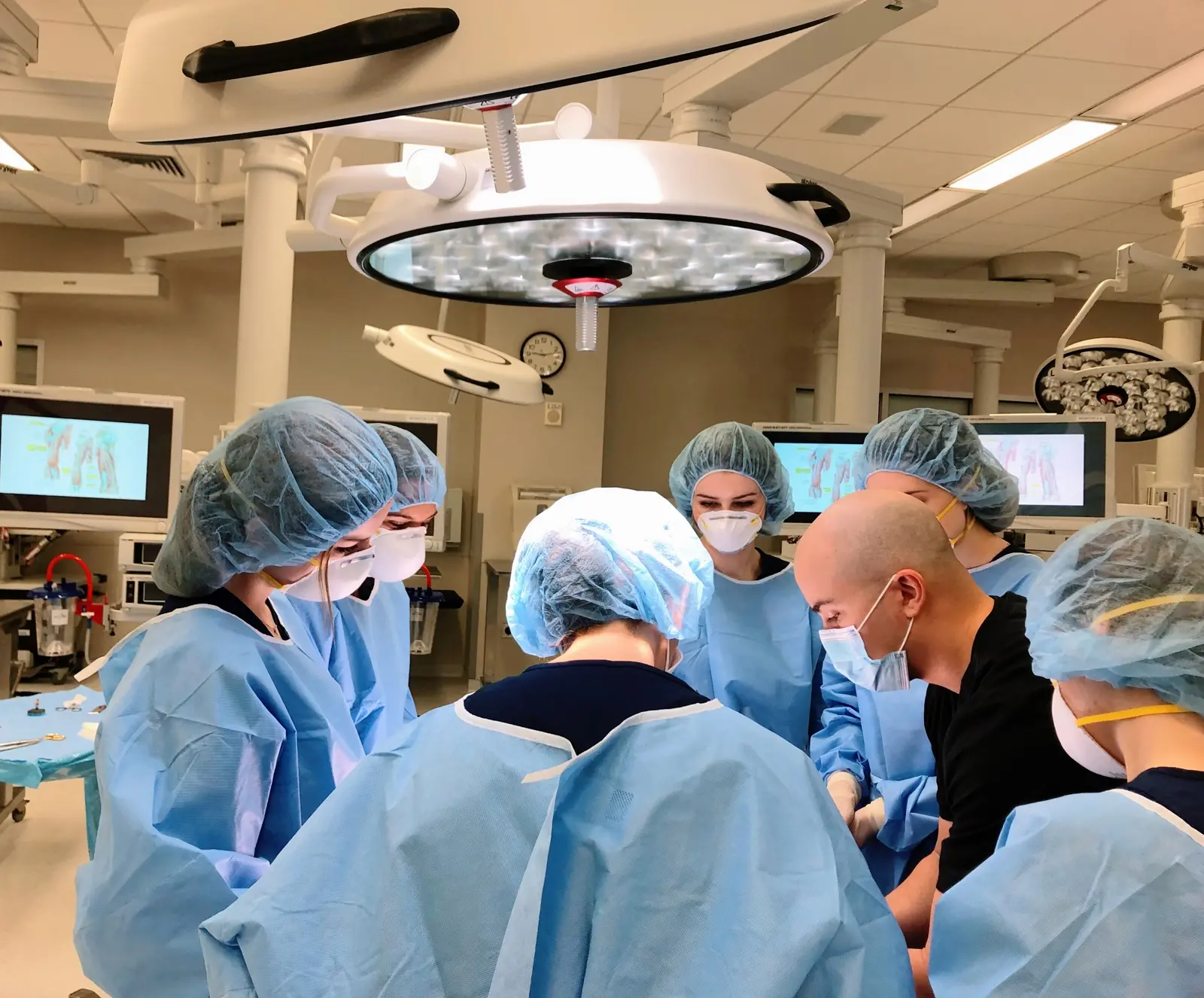 CAMLS A group of healthcare professionals wearing blue surgical gowns, masks, and hairnets are gathered around an operating table under a large surgical light in a modern operating room. They appear to be performing a medical procedure.