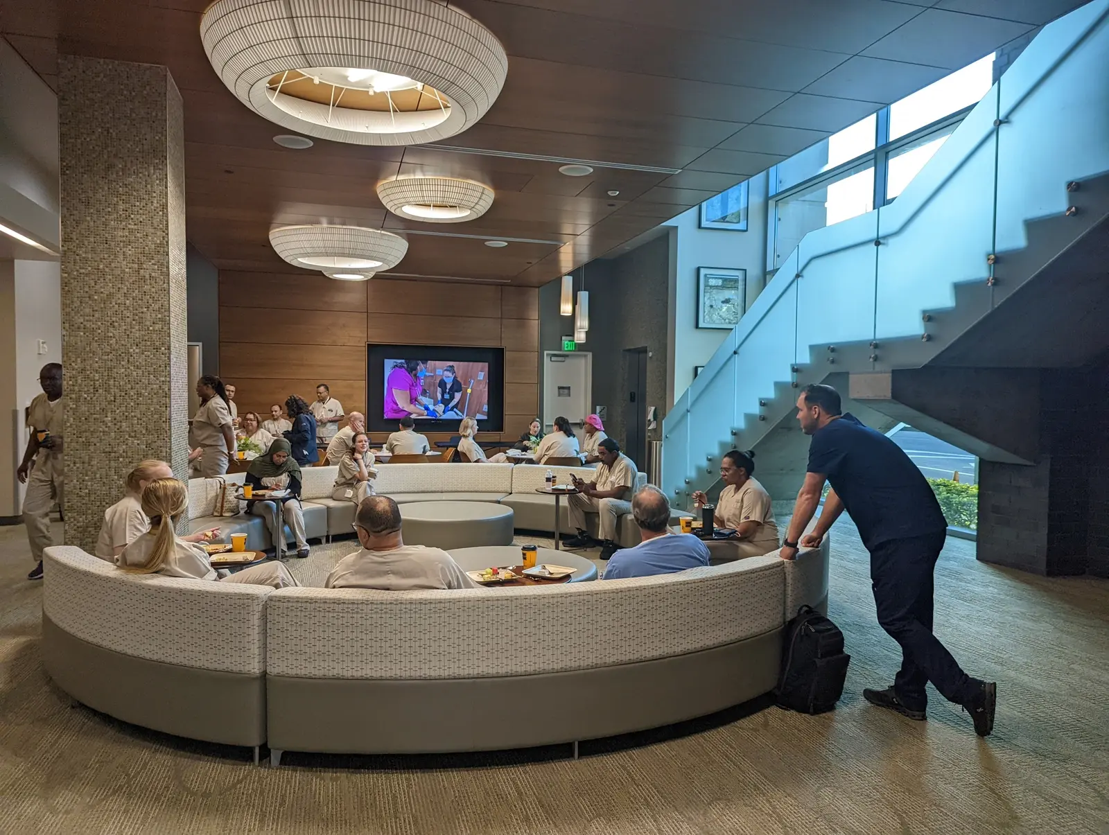 CAMLS A group of people in white uniforms sits and stands around a curved, modern sofa, watching a large TV screen in a spacious, well-lit lounge area. A man in dark clothing stands by a staircase on the right. The room features contemporary decor and large, circular ceiling lights.