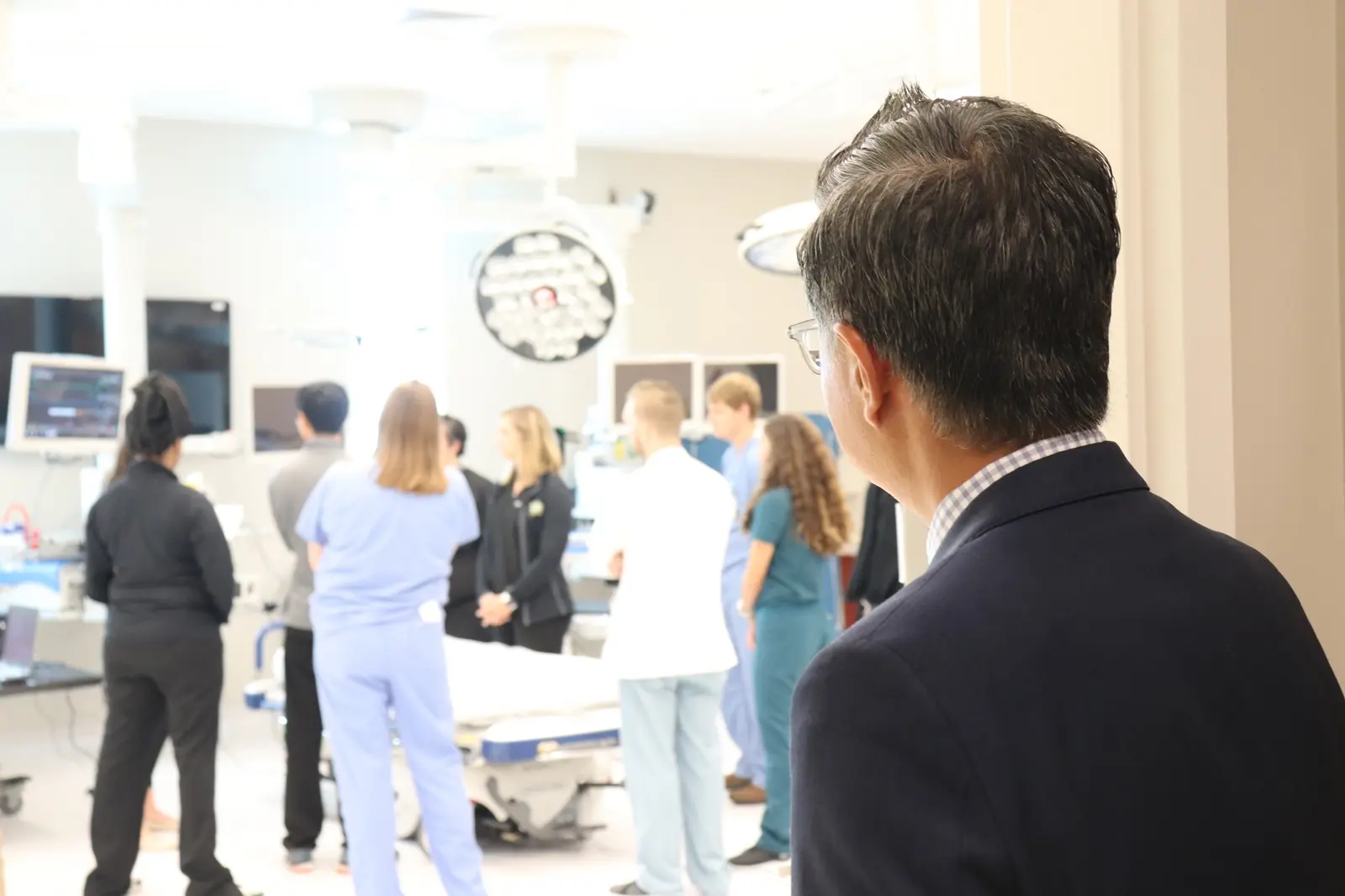 CAMLS A man in a suit stands in the foreground, looking into a bright room where a group of medical professionals in scrubs and lab coats are gathered. The room is equipped with medical equipment and monitors, suggesting a hospital or medical facility setting.