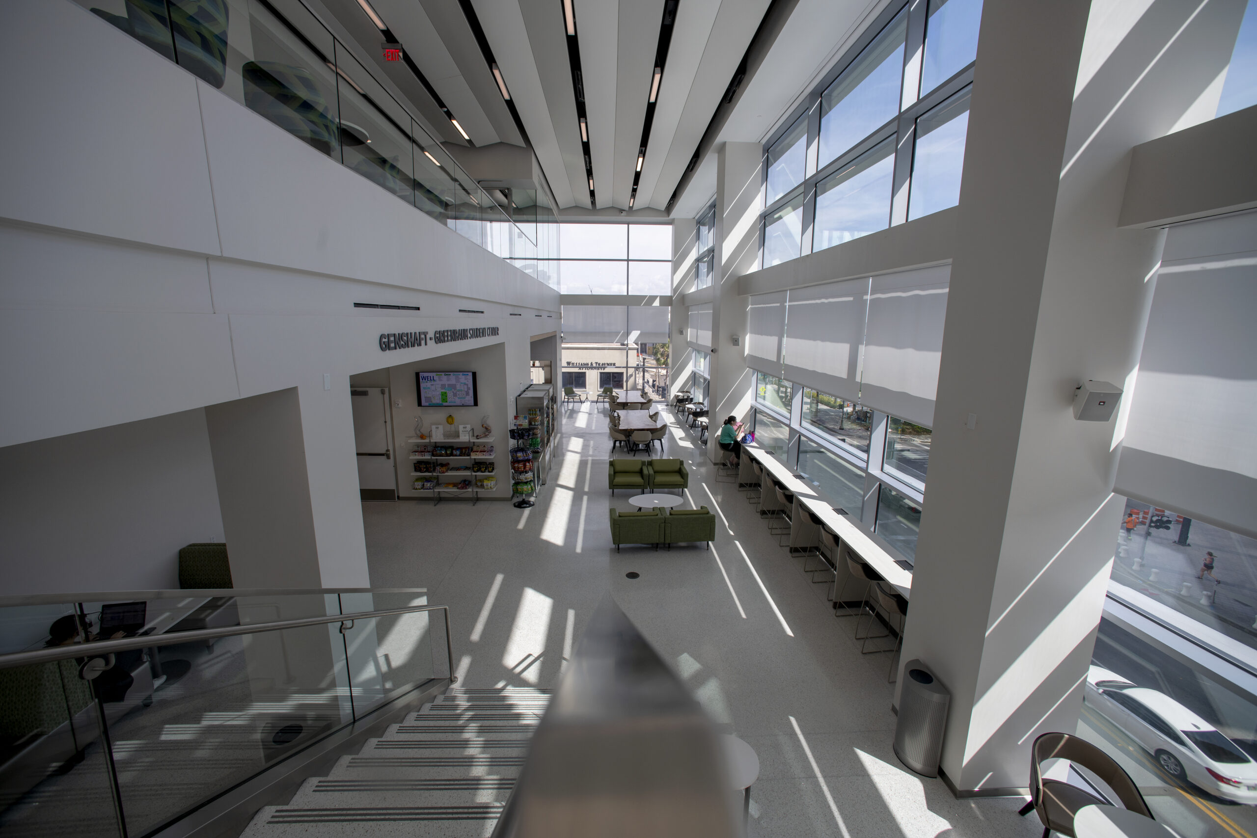 Wide-angle shot of a spacious, modern interior with large windows allowing ample natural light. The area features green furniture, a staircase with a glass railing, white walls, and a long table lined with chairs along the window. Shadows form geometric patterns.