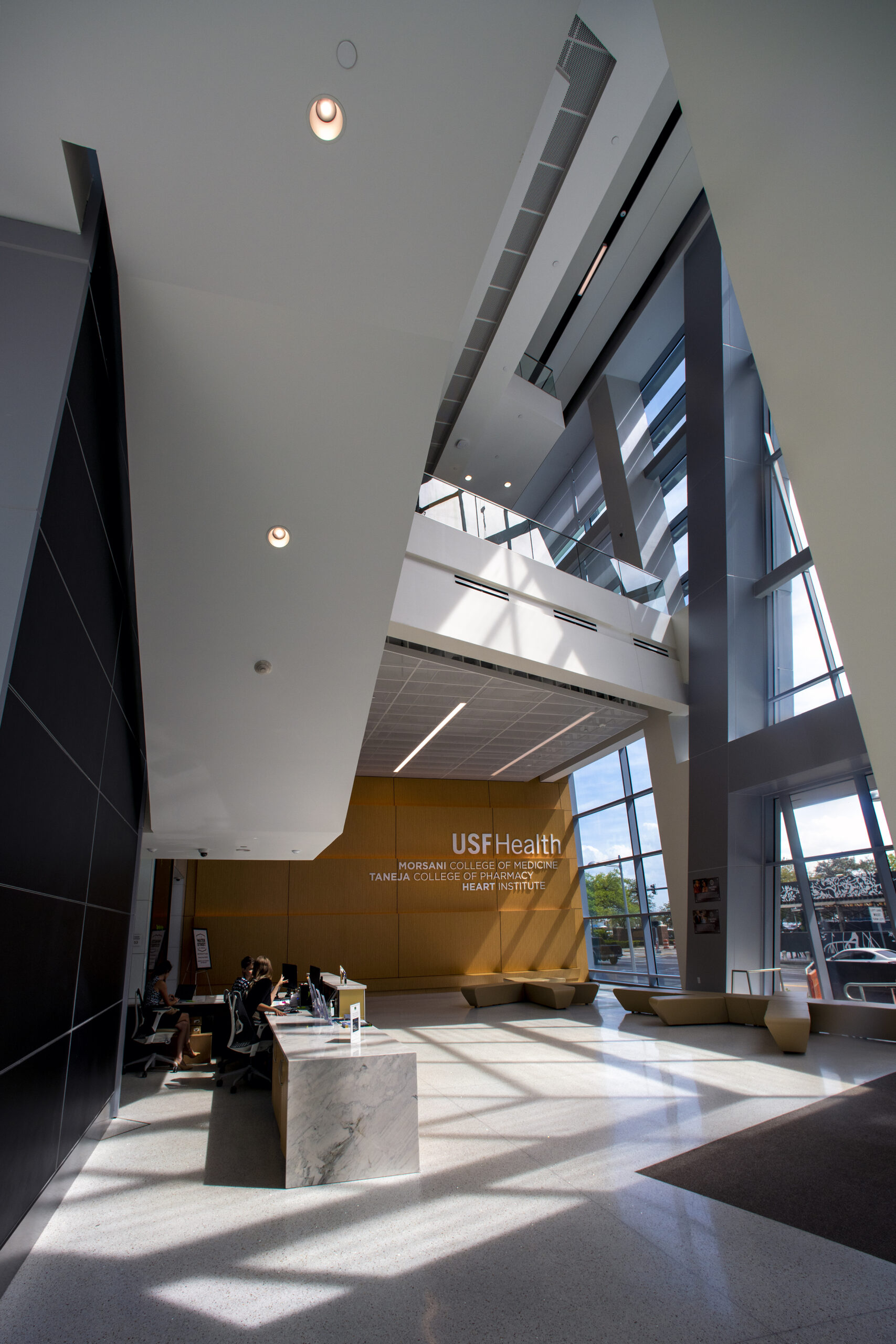 Modern lobby of USF Health Heart Institute featuring high ceilings, floor-to-ceiling windows, and sunlight streaming in. Reception desk with a marble counter sits in the center, and seating areas are visible.