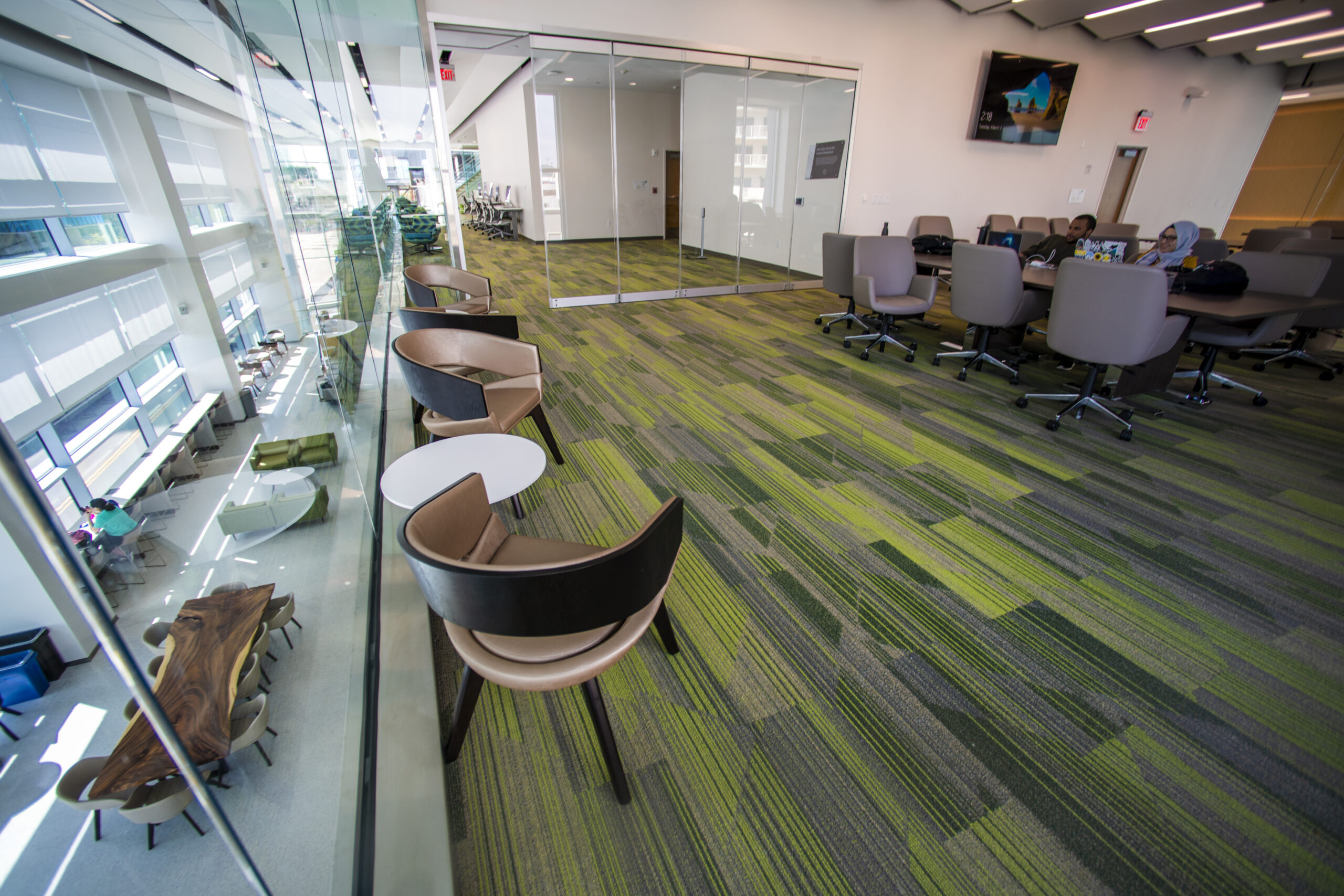 Spacious modern office with green-striped carpet, glass-walled conference room, and open seating area. Features a mix of chairs and tables, both individual and communal, with lots of natural light from large windows. Upper level overlooks a meeting space below.