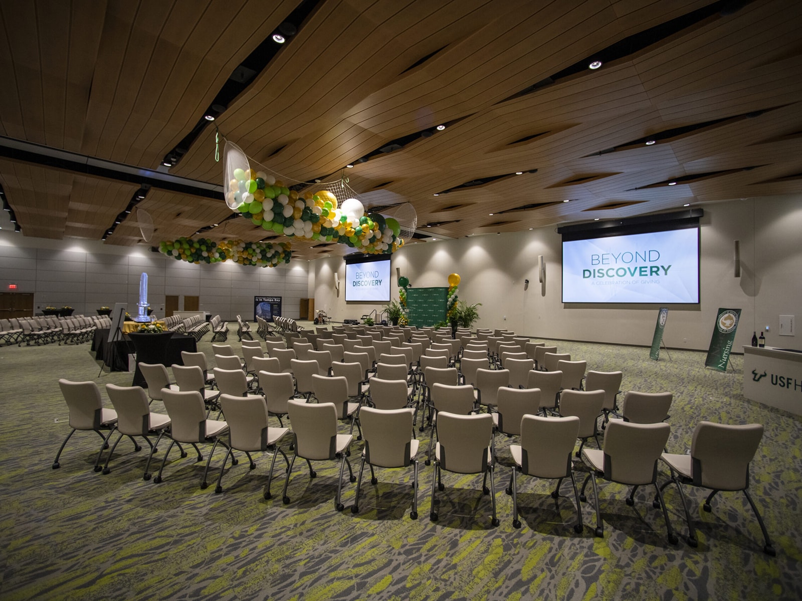 A spacious event hall with rows of empty chairs facing two large projection screens displaying "Beyond Discovery." The hall is decorated with green and yellow balloons. Tables with refreshments and displays are set up on the left side.