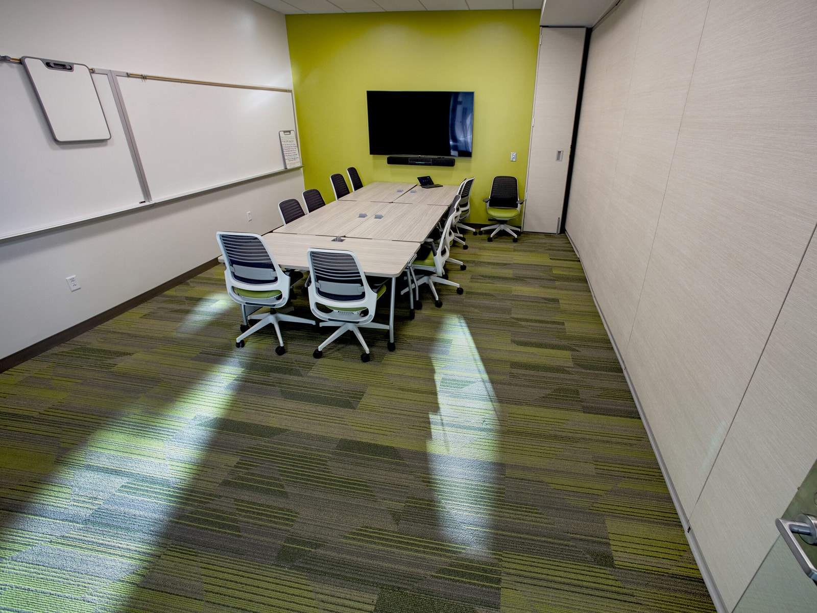 A modern conference room with lime green walls and a green striped carpet. There's a long rectangular table with six chairs arranged around it. A large flat-screen TV is mounted on the wall, and a whiteboard with a marker tray is positioned on another wall.