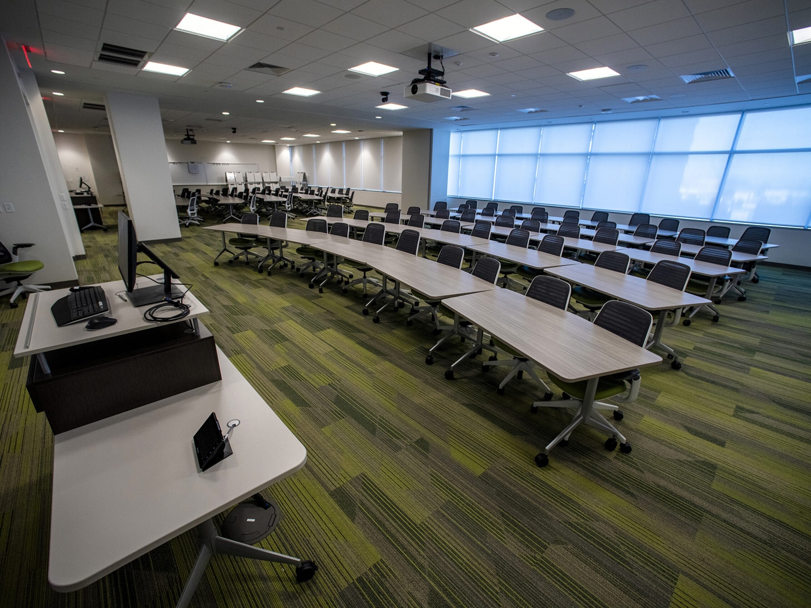 A modern classroom with rows of rectangular tables and chairs. The room features a combination of overhead fluorescent lights and natural light from large windows with blinds. A computer and equipment stand at the front on a teacher's desk. The carpet is green with a striped pattern.