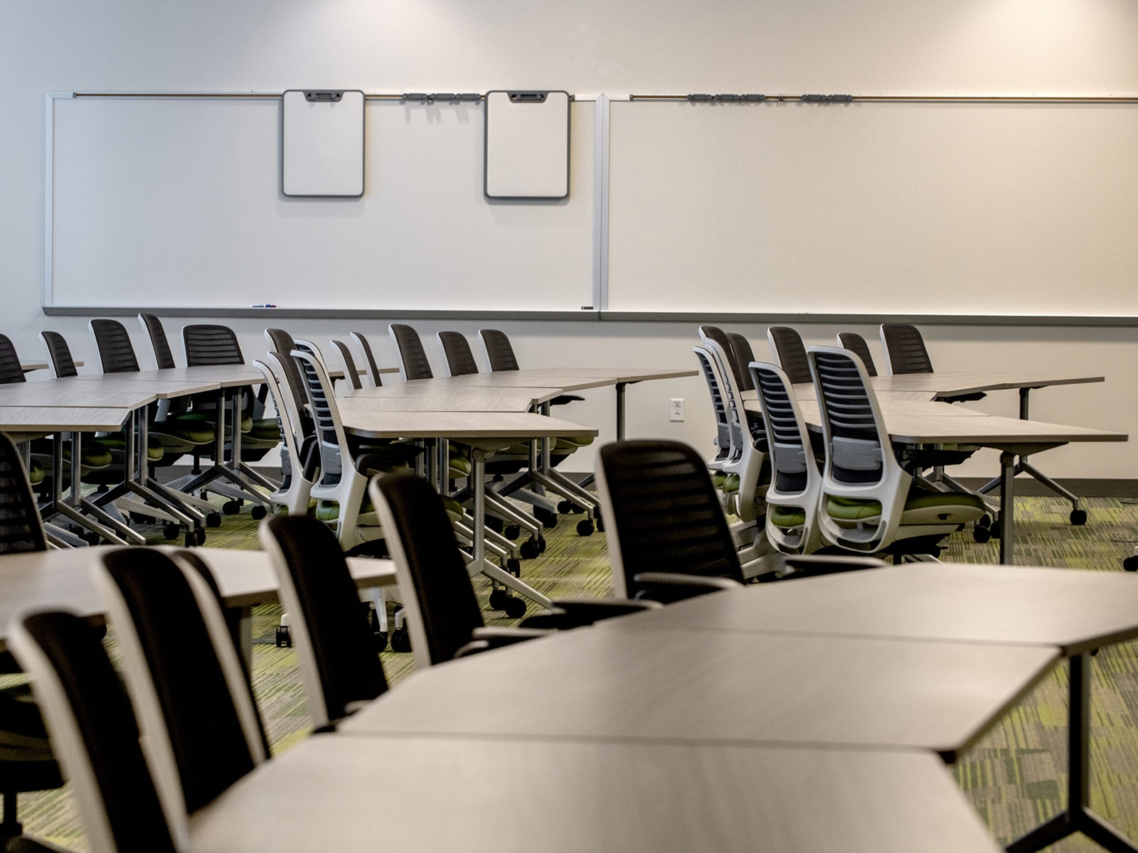 A modern classroom with multiple rectangular tables arranged in rows, each table surrounded by black chairs. The walls feature large whiteboards and two small whiteboards. The room is brightly lit with overhead lights.