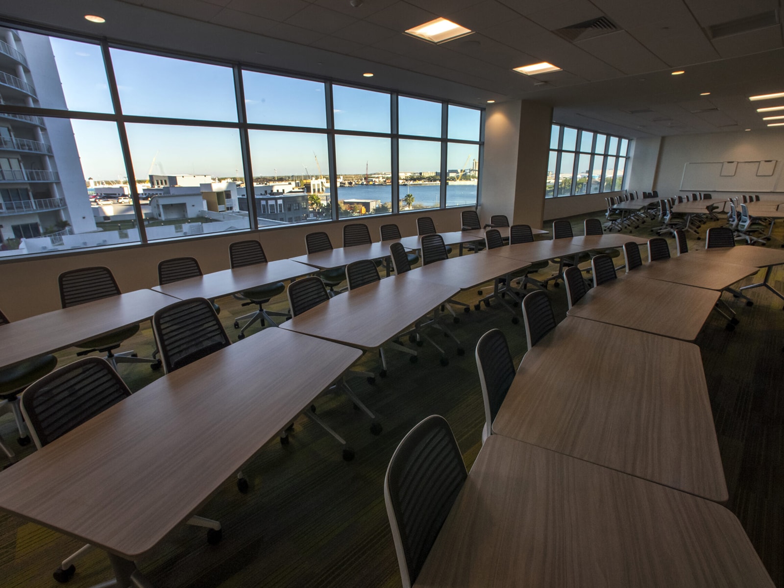 A modern conference room with long wooden tables and numerous ergonomic chairs. Large windows reveal an urban landscape with buildings and a shoreline in the distance. The room is well-lit by overhead lights and natural light from the windows.