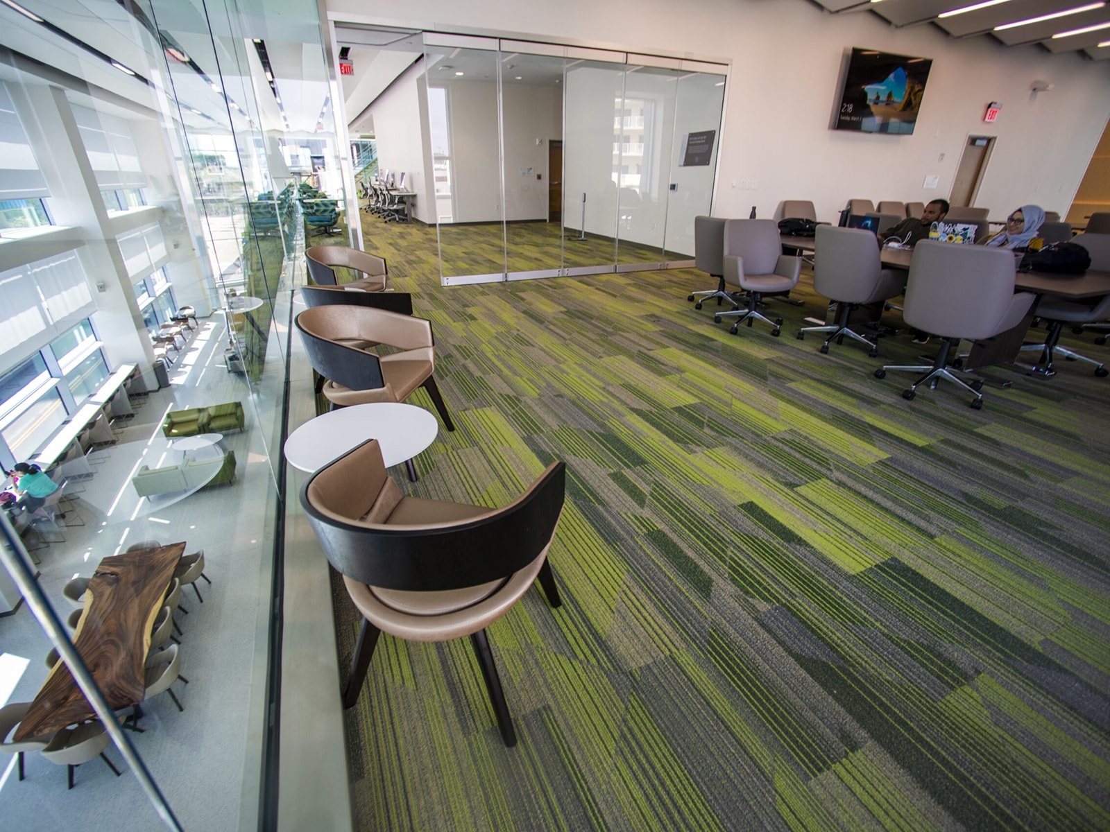 A modern office space with green and gray carpet, glass walls, and several seating areas. The foreground has round tables with chairs and lounge seating. The background features a conference room with a large table and chairs, separated by glass partitions.